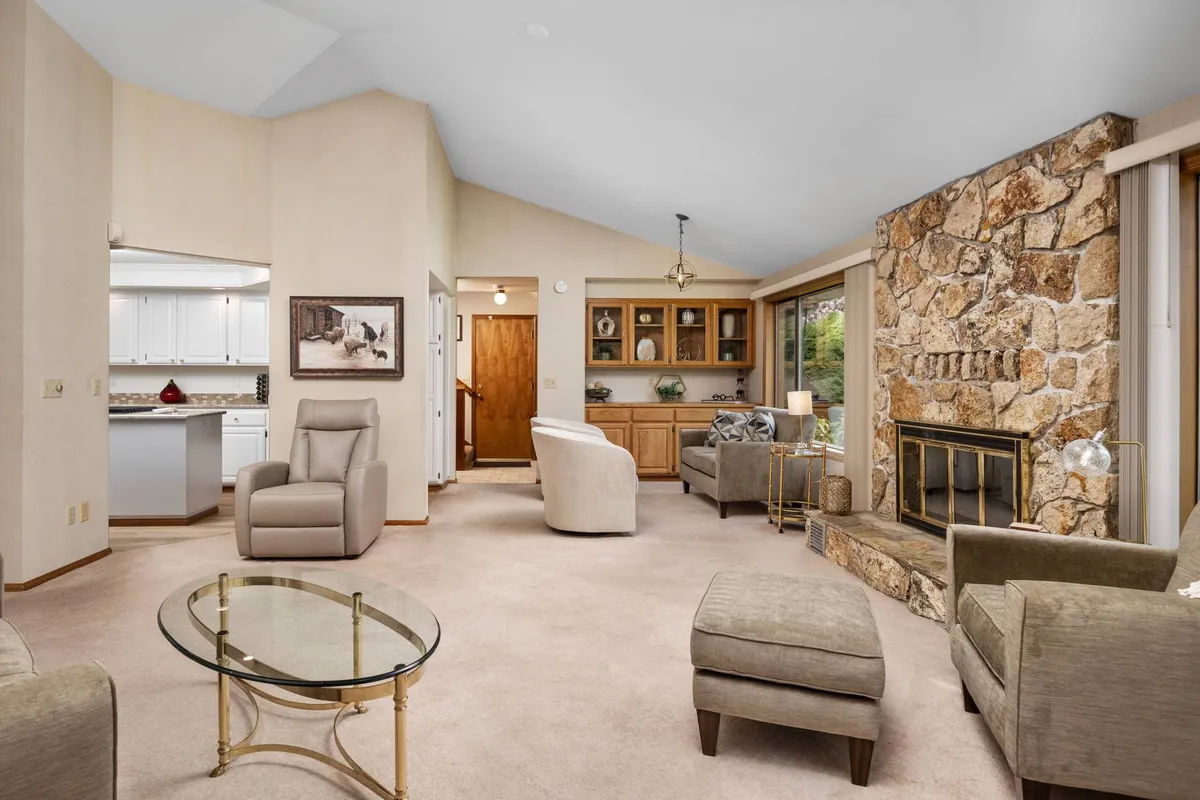 An inviting, sunlit living room featuring a stone fireplace, plush seating, large windows, and artwork. The layout is open and warmly decorated, ideal for both people and pets to unwind in a peaceful setting.