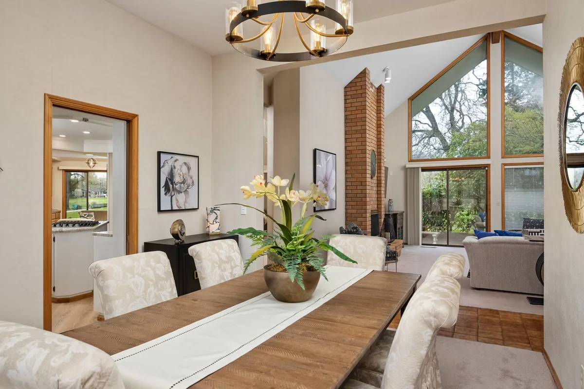 A formal dining room illuminated by a statement chandelier, with a long table set for gatherings, a brick fireplace adding warmth, and large windows bringing in natural light. A floral centerpiece completes the elegant setting.