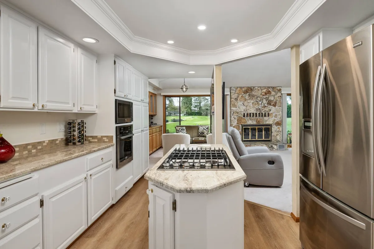 The image features a spacious, newly remodeled kitchen with a prominent island, a cozy stone fireplace, and expansive windows offering views of a lush garden. The layout encourages family interactions, with ample space for preparing meals, entertaining guests, and enjoying casual brunches or celebrations together. The warm lighting and open design invite multi-generational use.