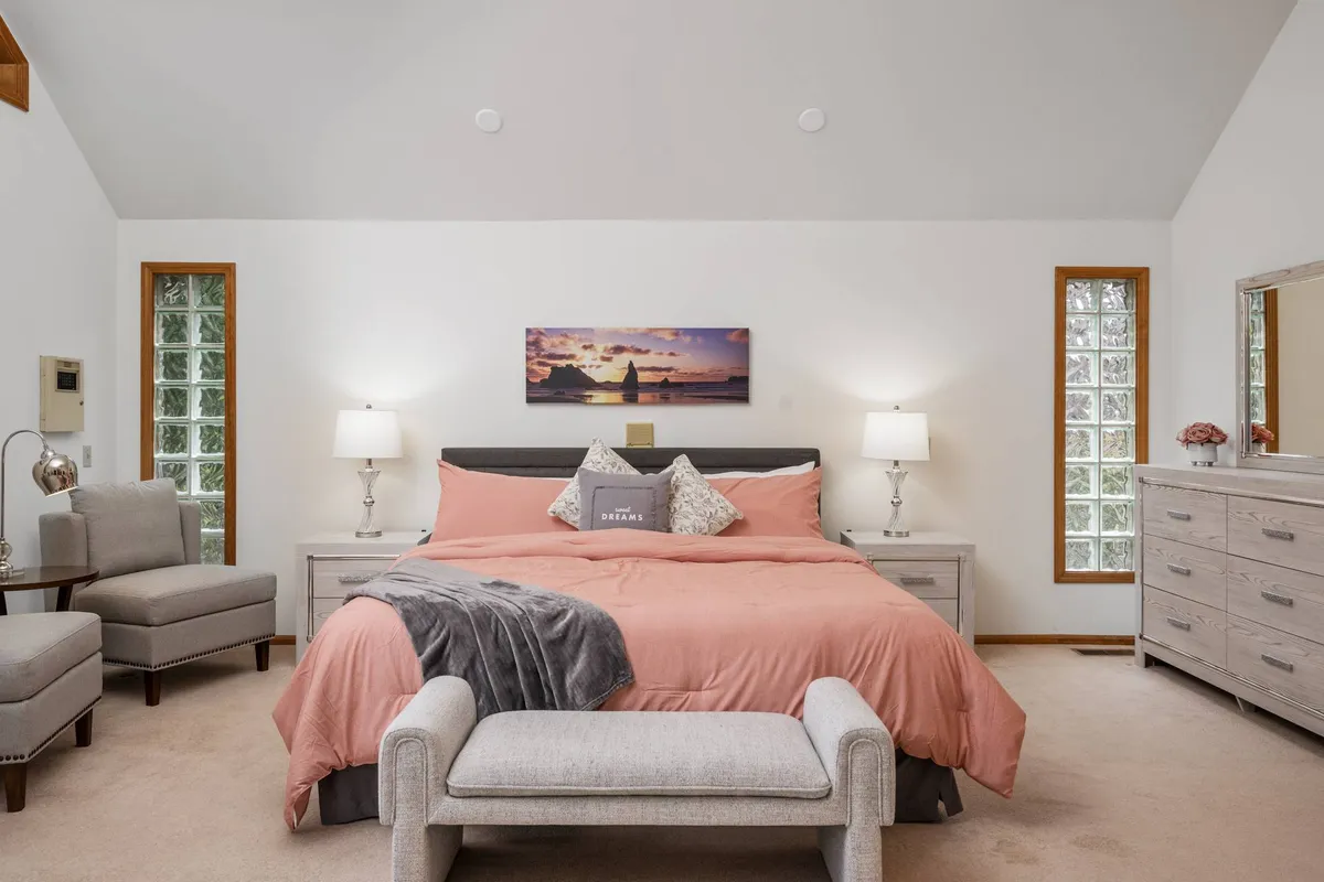 This image showcases a serene bedroom with a large bed dressed in coral bedding, a comfortable armchair in the corner, artwork above the bed, and a dresser with a mirror. Natural light filters into the room, creating a peaceful and inviting atmosphere.