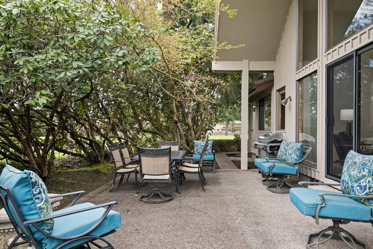 A backyard patio scene featuring a dining table, lounge chairs, a grill, and lush surrounding greenery. The setup encourages outdoor dining and relaxation, with ample seating positioned to enjoy the landscaped garden and natural views.
