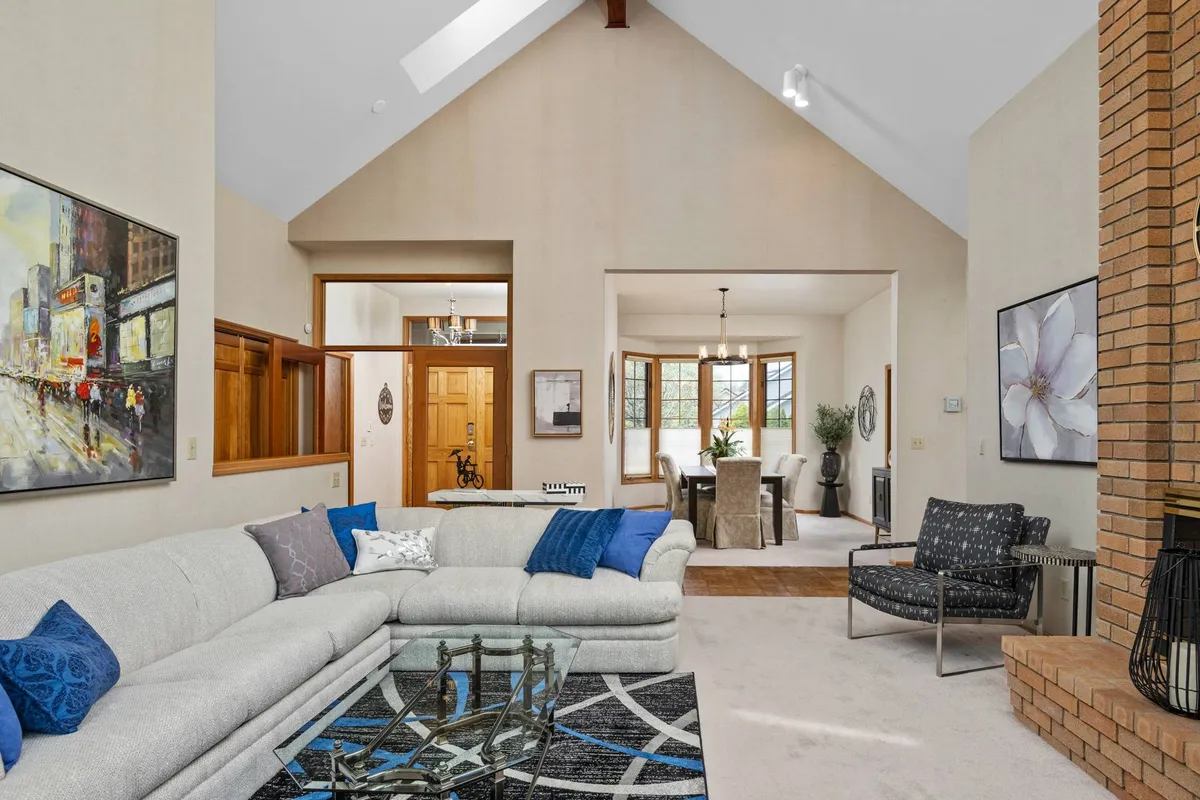 A spacious open-plan living and dining area featuring a large sectional sofa, brick fireplace, modern artwork on the walls, and a dining table set beside expansive windows flooding the space with natural light. The arrangement invites socializing, relaxation, and memorable gatherings.