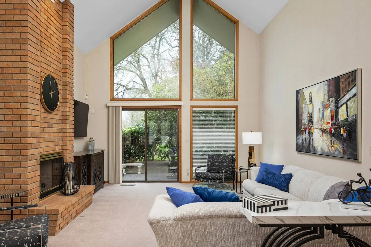 A cozy interior living space with a large brick fireplace, expansive windows letting in natural light, modern furniture, and tasteful artwork on the wall. The area appears open and comfortable, providing ample space for family and pets to relax together.