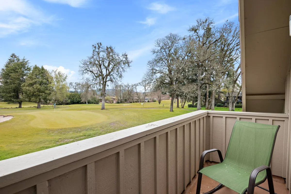 An upstairs balcony with a sleek railing overlooks the vibrant green expanse of the golf course. A green chair invites you to sit and enjoy the tranquil setting, surrounded by mature trees. The serene environment is ideal for soaking in sunsets or quietly reading while taking in panoramic views.