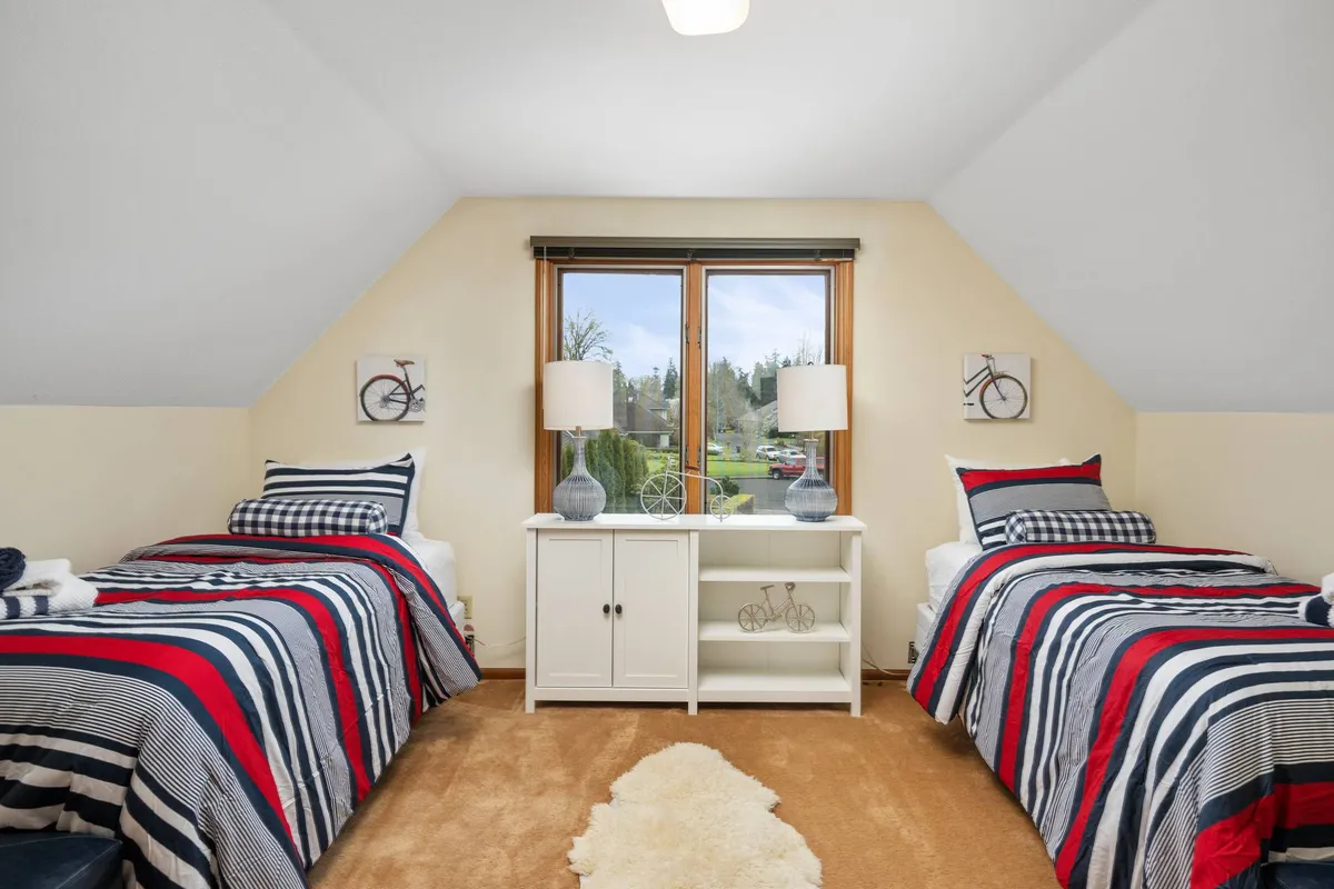 A cheerful bedroom featuring two beds with striped bedding, large windows that fill the room with natural light, decorative lamps, and a white shelving unit. The playful, friendly setup is ideal for children or friends.