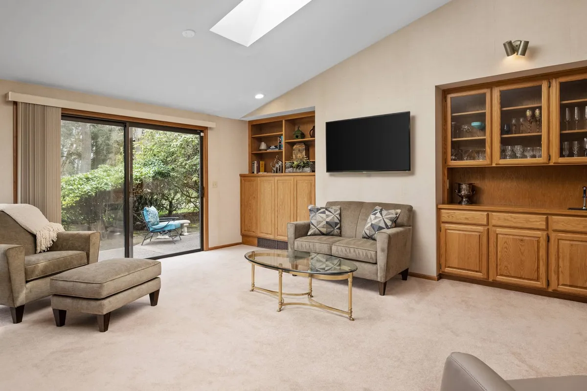 The image features a cozy seating area with a glass coffee table, comfortable sofa, bookshelves, and large windows that flood the space with natural light. The setup suggests a flexible family room ideal for both people and pets to unwind.