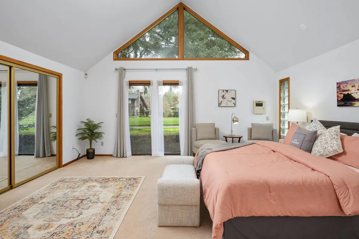 A peaceful bedroom with coral bedding, large sunlit windows, mirrored closet doors, and a comfortable seating area with chairs. The space is airy and reflective, suggesting both privacy and relaxation.